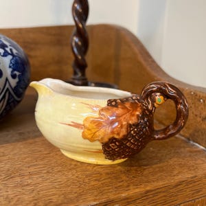 May include: A small, yellow ceramic creamer with a brown acorn-shaped handle and a decorative leaf design. The creamer has a spout for easy pouring and is set on a wooden surface. The handle features a small green accent.