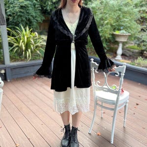 May include: A black velvet jacket with bell sleeves and lace trim, worn with a cream lace-trimmed dress. The outfit is completed with black boots. A white chair is in the background.