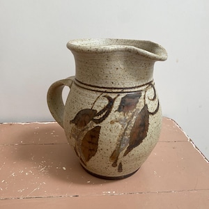 May include: A speckled beige ceramic jug with a handle and a pouring spout. The jug is decorated with brown leaf designs and a dark brown band around the middle. The jug sits on a pink surface.