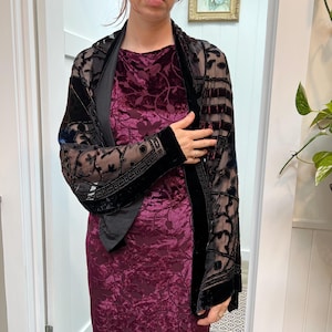 Puede incluir: Un vestido de terciopelo burdeos se combina con una chaqueta negra transparente. La chaqueta presenta intrincados diseños florales y ribetes de terciopelo. Las mangas tienen un borde decorativo. El conjunto es adecuado para ocasiones formales.