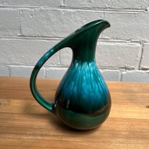 May include: A vintage ceramic jug with a distinctive handle and a striking colour gradient. The jug transitions from a deep forest green at the base to a vibrant teal near the top, creating a visually appealing effect. The handle is curved and adds to the jug's elegant design.