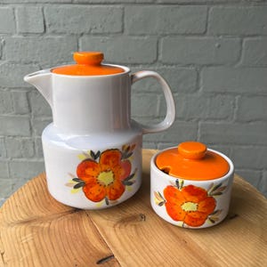 Puede incluir: Un juego de cafetera y azucarero de cerámica blanca con tapas naranjas. El juego presenta un diseño floral con flores naranjas y amarillas y hojas verdes. La cafetera tiene un asa y un pico.