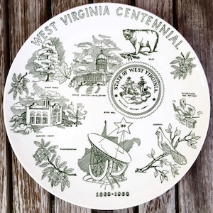 May include: A white ceramic plate with a green design commemorating the West Virginia Centennial. The plate features illustrations of state symbols, including a sugar maple tree, the state capitol building, a bear, a cardinal, and a radio telescope. The text "West Virginia Centennial 1863-1963" is printed on the plate.