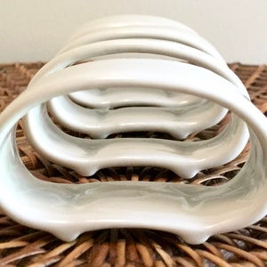 May include: A stack of four white ceramic napkin holders. Each holder has a curved, open design with a smooth, glossy finish. The holders are arranged on a woven, brown surface.