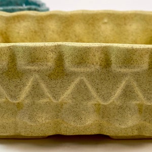 May include: A square, pale yellow ceramic planter with a textured, geometric design. The planter has a slightly scalloped rim and a wide, open top.