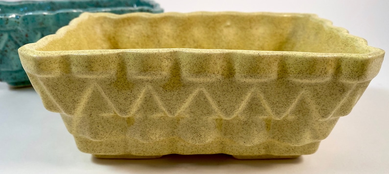 May include: A square, pale yellow ceramic planter with a textured, geometric design. The planter has a slightly scalloped rim and a wide, open top.