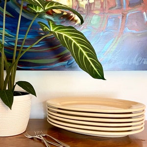 May include: A stack of six cream-colored oval plates. The plates are stacked on a wooden table with two silver forks in front of them.