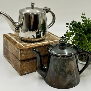 May include: Two stainless steel teapots, one shiny and the other tarnished, sit on a decorative wooden box. The teapots have spouts, handles, and lids. A small green plant is in the background.