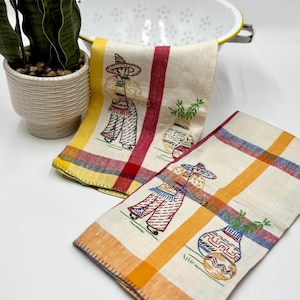 May include: Two cream-colored embroidered kitchen towels with a Mexican theme. Each towel features a figure in a sombrero and a vase with plants. The towels have red, yellow, and orange plaid borders. A white colander with a yellow rim is in the background.