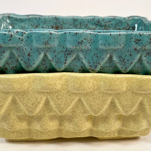 May include: Two square ceramic planters, one teal with brown speckles and one yellow with brown speckles. Both planters have a geometric design around the top edge.