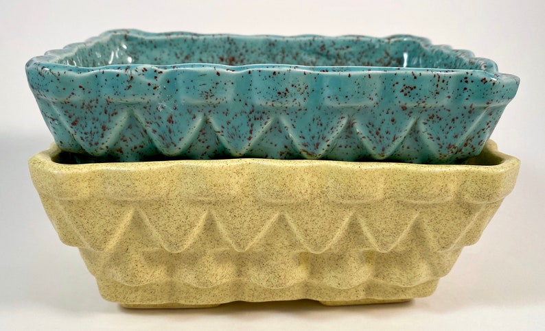 May include: Two square ceramic planters, one teal with brown speckles and one yellow with brown speckles. Both planters have a geometric design around the top edge.