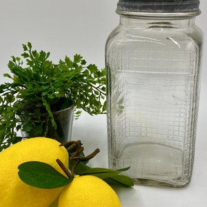 May include: A clear glass jar with a gray metal lid. The jar has a textured surface and is empty. Two yellow lemons with green leaves are in the foreground.