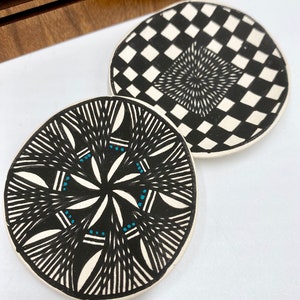 May include: Two round ceramic coasters with black and white geometric designs. One coaster features a checkerboard pattern with a black and white swirl in the center. The other coaster has a black and white geometric design with blue accents.