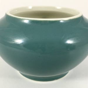 May include: A small, round, teal ceramic pot with a white rim. The pot has a smooth, glossy finish.