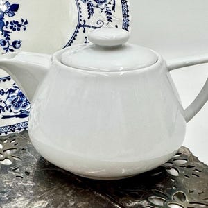 May include: A white ceramic teapot with a lid and a handle. The teapot is round with a spout and sits on a decorative metal trivet. A blue and white floral patterned plate is in the background.