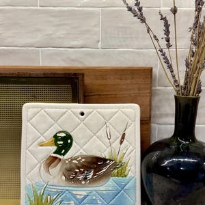 May include: A square ceramic wall plaque featuring a raised relief of a mallard duck swimming in blue water with green reeds and cattails. The background has a white quilted pattern with a small hole at the top center for hanging purposes.
