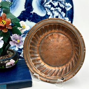 May include: A copper-colored, round decorative plate with a detailed, embossed floral design in the center. The plate has a ribbed edge and is displayed on a clear stand. A blue and white patterned plate is in the background.
