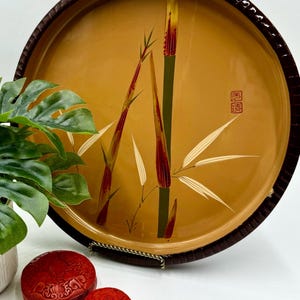 May include: A round, brown wooden tray with a raised rim and a painted design of bamboo stalks in shades of green, red, and yellow. The tray has a brown border and a red stamp with Chinese characters.