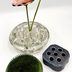 May include: A clear glass flower frog with multiple circular openings holding two pink flowers, a round green metal pin frog with sharp needles, and a small square blue ceramic flower frog with seven circular holes for floral arrangements.