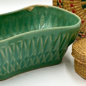 May include: An assortment of decorative items. A rectangular, seafoam green ceramic planter with a textured diamond pattern. A woven basket with a lid and a round, woven basket with red and green accents.