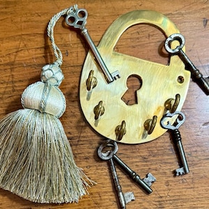Puede incluir: Un porta llaves de latón con un diseño de candado grande y cinco ganchos para llaves. El porta llaves está decorado con un borla y varias llaves antiguas.