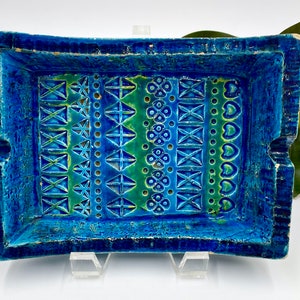 May include: Rectangular ceramic tray with a textured, dark blue exterior and a patterned interior. The interior features rows of geometric designs in shades of blue and green. The tray is supported by a clear stand.