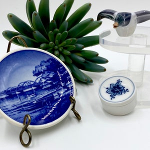 May include: A blue and white porcelain plate with a depiction of a bridge and a boat. The plate is on a metal stand. There is also a small blue and white porcelain box with a floral design.