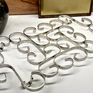 May include: A rectangular silver-toned metal trivet featuring a repeating pattern of interconnected scrollwork loops. The design consists of multiple curved metal bands joined together to form a flat, open-work structure with a polished finish.