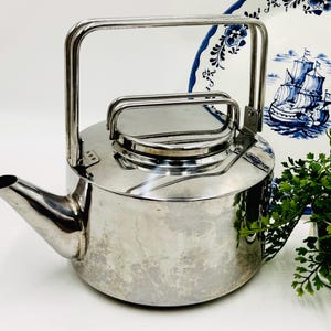 May include: A polished stainless steel kettle with a cylindrical body, a short angled spout, and a dual-handle design. The main handle is tall and rectangular, while a smaller handle is attached directly to the circular lid on top.