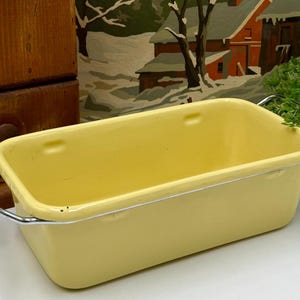 May include: A rectangular, pale yellow enamelware baking dish with silver-colored handles. The vintage kitchenware has a slightly rounded rim and is empty. The background includes a wooden cabinet and a painting of a winter scene.