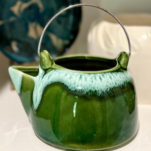 May include: A vintage ceramic pitcher with a dark green glaze and a light green, textured rim. It has a silver metal handle and a spout. The pitcher is likely used for serving beverages or as a decorative item.