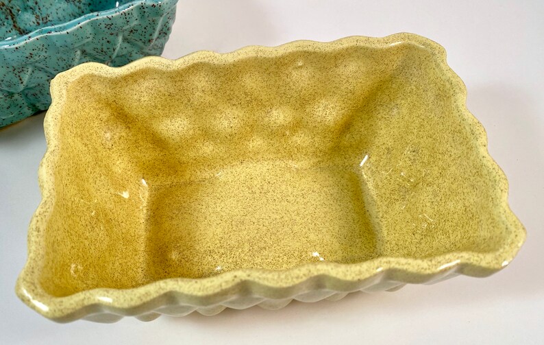 May include: A yellow ceramic planter with a textured, speckled finish. The planter has a square shape with rounded edges and a slightly wavy rim.
