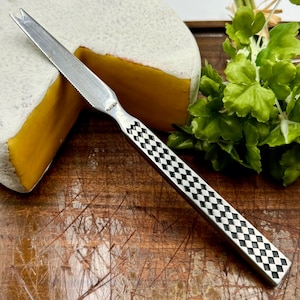May include: A stainless steel cheese knife with a checkered black and white handle. The knife is positioned next to a wheel of cheese with a yellow interior and a white rind. Fresh green herbs are in the background.