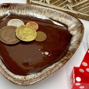 May include: A brown ceramic dish with a white rim and a glazed finish. The dish is shaped like a triangle and has five coins inside. The coins are all different sizes and have a gold color.