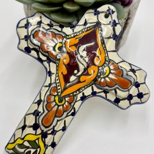 May include: A white ceramic cross with a blue, yellow, orange, and red floral design. The cross has a lattice pattern and is decorated with intricate details.