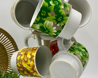 May include: A white, distressed-look mug tree holds six floral-patterned mugs. The mugs feature designs in green, yellow, and red. The tree has a round base and a decorative top. A gold-colored woven plate and a small potted plant are in the background.