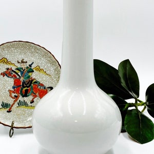 May include: A white ceramic vase with a tall, slender neck and a rounded body. The vase is simple and elegant.