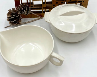May include: Two white ceramic bowls, one with a lid. The bowl with the lid has a small handle on each side. The other bowl has a single handle on the side. Both bowls are empty.