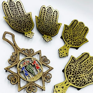 May include: Five gold-colored metal ornaments with intricate designs. Four of the ornaments are shaped like hands with the word "ISRAEL" written on the back. The fifth ornament is a six-pointed star with a colorful image of three figures inside.