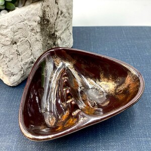 May include: A brown and gold ceramic ashtray with a triangular shape and a unique design. The ashtray has four indentations for cigarettes.