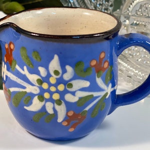 May include: A small blue ceramic creamer with a white floral design. The creamer has a brown rim and a handle.