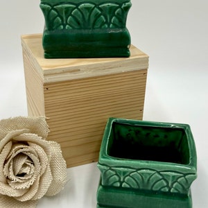 May include: Two green ceramic square planters with a decorative design around the top edge. The planters are sitting on a wooden surface.