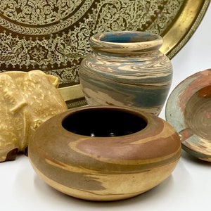 May include: A collection of decorative pottery items, including a camel figurine, a small vase, a round bowl, and an ashtray. The pieces feature earthy tones of brown, beige, and blue, with a brass tray in the background. The items are suitable for home decor.