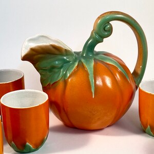 May include: A vintage orange and green ceramic pumpkin-shaped pitcher with a handle and four matching cups. The pitcher has a detailed leaf design.