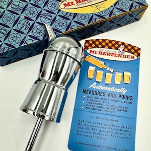 May include: A silver-colored Mr. Bartender automatic liquor dispenser with a long, thin spout. The dispenser is designed to measure and pour one or many drinks from a single tilt. The text on the card reads "The Famous Mr. Bartender Automatically Measures and Pours".