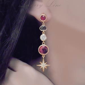 Peut inclure: Boucles d'oreilles pendantes dorées avec un motif d'étoile. Les boucles d'oreilles présentent une série de pierres précieuses colorées, dont des pierres rouges, noires et claires. Elles font partie de la ligne Azaliya Luxury.