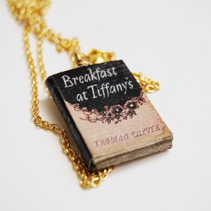 May include: A gold chain necklace with a miniature book pendant. The book cover is black with white text that reads "Breakfast at Tiffany's" and the author's name, Truman Capote.
