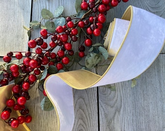 May include: A festive arrangement featuring a white ribbon with gold trim, alongside a branch of red berries and green leaves. The ribbon is partially rolled, creating a decorative element. The image is set against a wooden background.