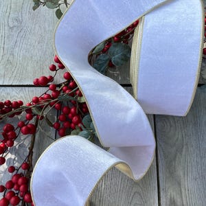 May include: A wide, white velvet ribbon with a metallic gold edge, coiled and draped over a decorative branch featuring numerous small, round, bright red berries and muted green leaves, creating a high-contrast seasonal display.