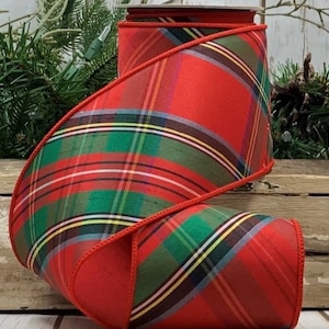 May include: A roll of wired ribbon featuring a classic plaid pattern in shades of red, green, yellow, and navy blue. The fabric has a smooth, slightly lustrous finish with a solid red stitched edge running along both sides of the ribbon.
